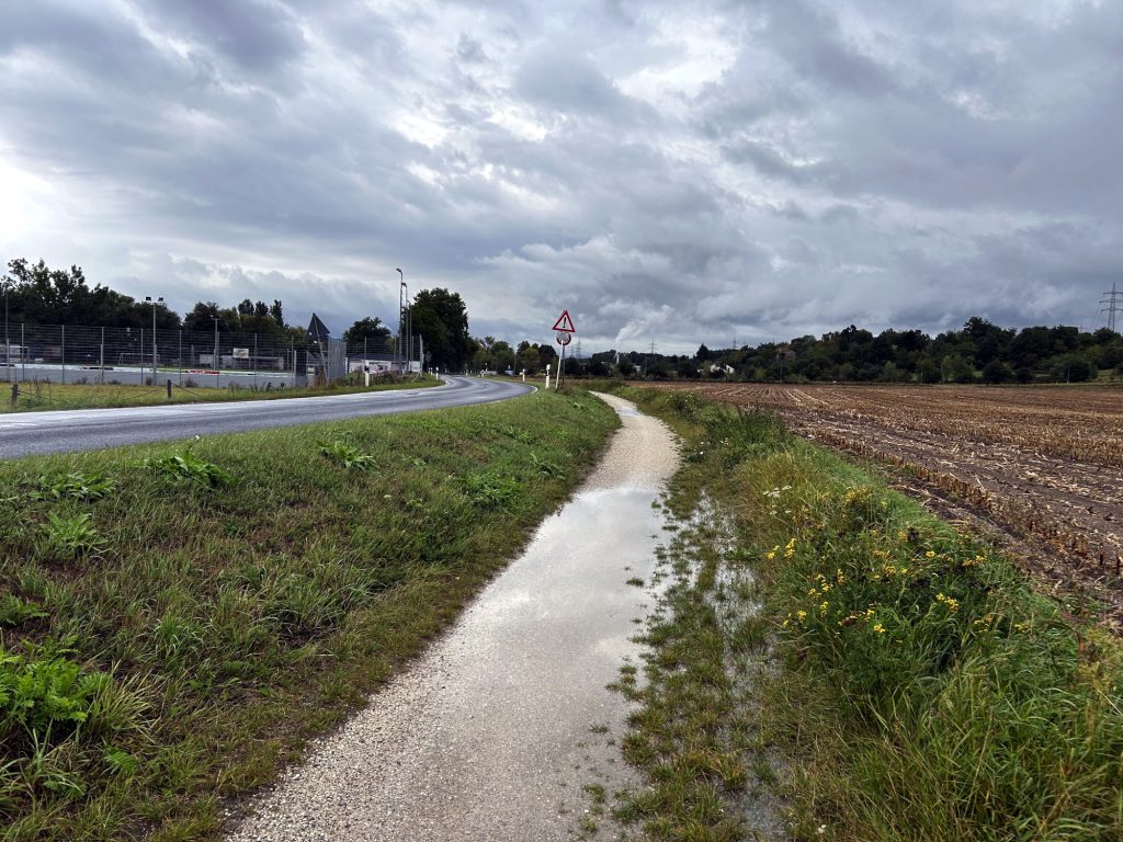 vLnR: 
K 63 mit geteerte Oberfläche
Böschung mit kurzem Bewuchs, nach rechts abfallend
Feldweg mit Schotter. Mehrere große Pfützen sichtbar.
Böschung mit mittlerem Bewuchs, nach links abfallend
Abgeerntetes Feld