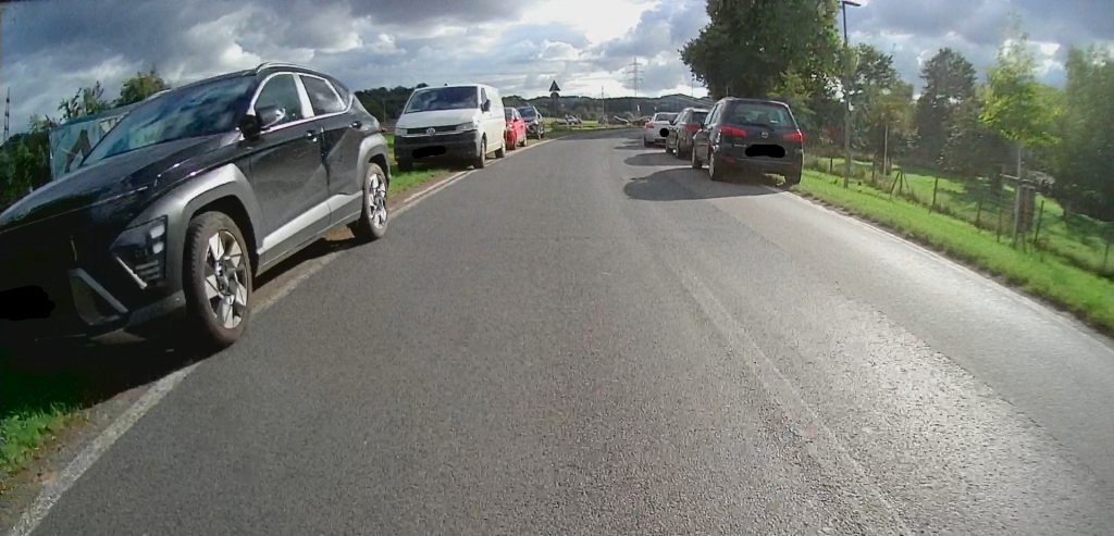 Straße am Sportplatz Miesenheim. Links eine Reihe parkender Fahrzeuge neben dem Asphalt. Rechts ebenso, aber teils auf dem Fahrstreifen stehend und diesen Blockierend
