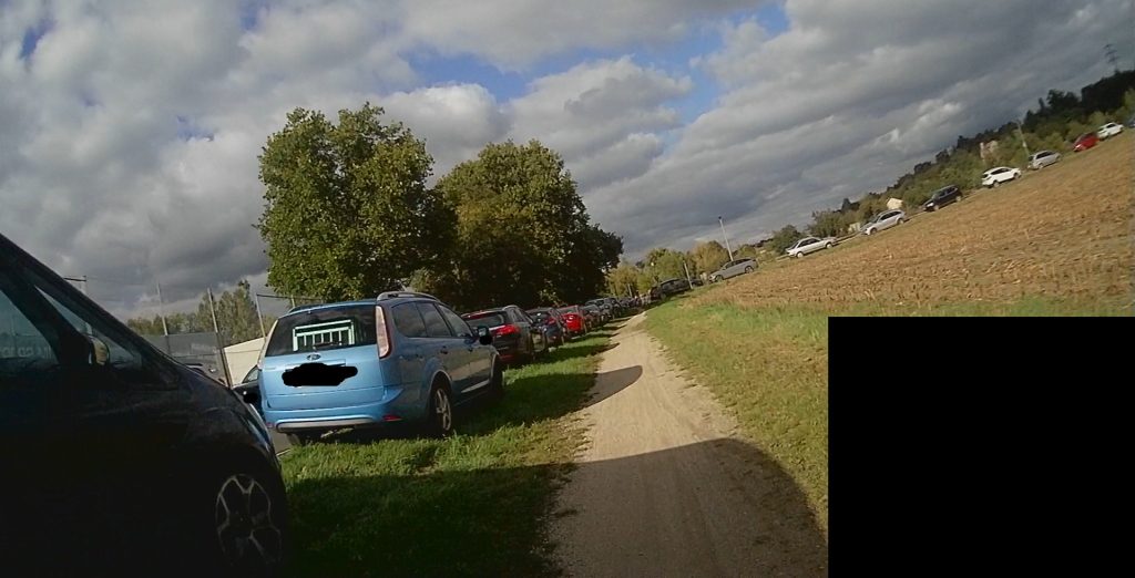 Bestehender Feldweg am Sportplatz Miesenheim. Links eine Reihe parkender Autos im Grünen. Dahinter Teilweise Straße mit weiteren parkenden Autos auf der anderen Straßenseite erkennbar. Grade Blick auf Zufahrt Schützenhaus. Auch dort gesamter Weg mit parkenden PKW belegt.